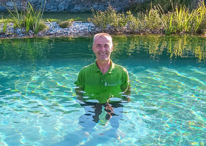 Zu sehen ist ein Mann, der am Rand eines Schwimmteiches sitzt. Einen Fuß hält er im Wasser. Hinter ihm erkennt man einen großen Schwimmteich sowie viele Pflanzen.