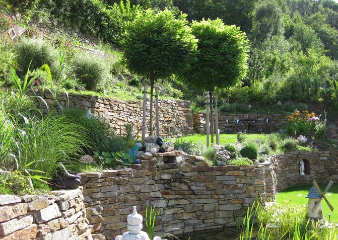Ein Hang mit vielen Grünpflanzen wird durch viele Steinmauern terrassiert. So entstehen auch kleine Wiesenflächen.