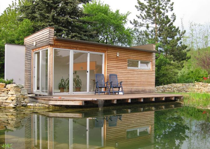 Ein kleines Holzhaus mit Holzsteg Richtung Schwimmteich. Am Holzsteg stehen 2 Gartenmöbel.