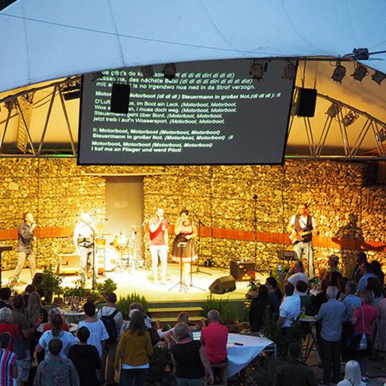 Im Hintergrund sieht man eine Bühne auf der eine Band mit fünf Personen spielt. Im Vordergrund, vor der Bühne, steht das Publikum bei Stehtischen und lauscht der Musik. Oberhalb der Bühne hängt eine große Leinwand auf der die Liedtexte zum Mitsingen stehen.