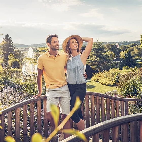 Eine Frau und ein Mann stehen mittig im Bild auf einer kleinen Holzbrücke. Im Hintergrund sieht man einen großen Garten und einen Springbrunnen.