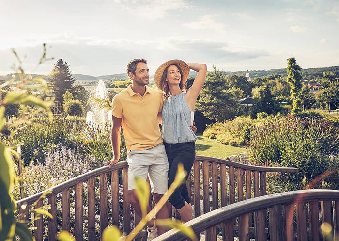 Eine Frau und ein Mann stehen mittig im Bild auf einer kleinen Holzbrücke. Im Hintergrund sieht man einen großen Garten und einen Springbrunnen.