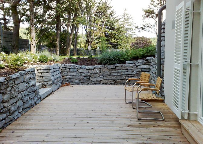 Eine tieferlegte Terrasse mit Holzbretter, die mit einer niedrigen Steinmauer vom Garten getrennt ist.