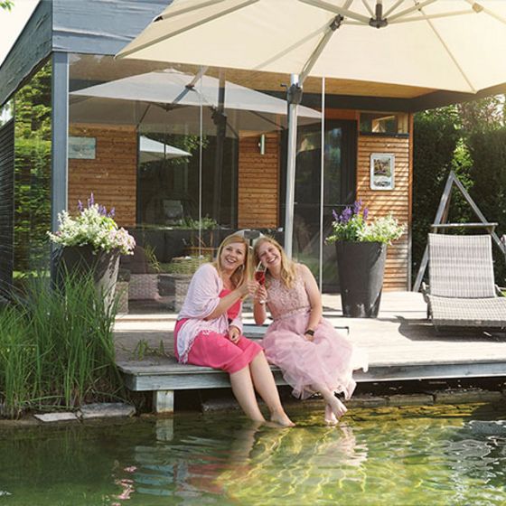 Zwei Frauen sitzen auf einem Steg bei einem Schwimmteich. Sie halten die Füße in das Wasser und stoßen mit Sektgläsern an. Im Hintergrund stehen Liegen und oben im Bild ist ein Sonnenschirm zu sehen. 