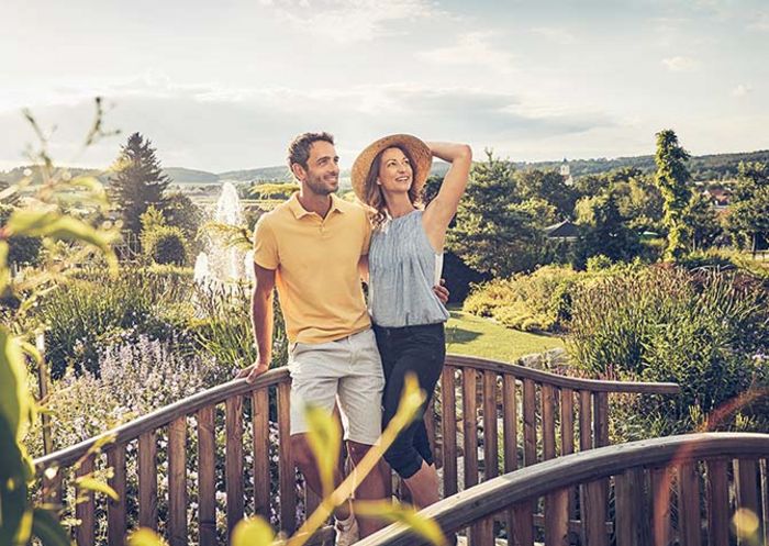 Ein Mann und eine Frau stehen auf einer kleinen Holzbrücke. Im Hintergrund sieht man einen großen Garten und einen Springbrunnen.